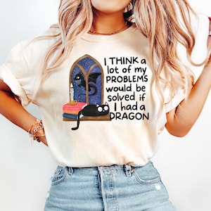May include: Cream-colored t-shirt with the text "I THINK a lot of my PROBLEMS would be solved if I had a DRAGON." The graphic features a black cat, a dragon, a candle, and books. The design is whimsical and fantasy-themed.