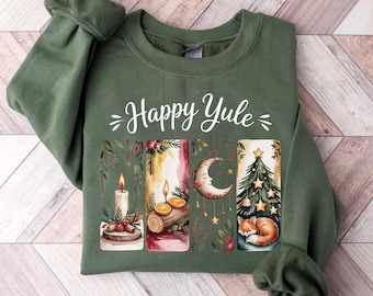 May include: A green sweatshirt with the words "Happy Yule" in white script. The graphic features watercolor illustrations of candles, a crescent moon, and a Christmas tree with a sleeping fox. The design has a festive, holiday theme.