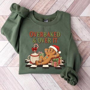 May include: Green crewneck sweatshirt with the text "OVERBAKED & OVER IT" in red and green. The graphic features a gingerbread man wearing a Santa hat, a mug of hot chocolate, and wrapped gifts. The sweatshirt has cuffed sleeves.