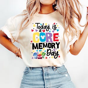May include: Cream-colored t-shirt with the text "Today is a CORE MEMORY Day" in colorful letters. The design includes Mickey Mouse ears and characters from the movie Inside Out. The shirt is paired with light blue jeans.