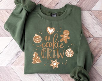May include: A dark green sweatshirt with the text "The Cookie Crew" in a cursive font, along with gingerbread-themed illustrations. The design includes a gingerbread man, heart, star, Christmas tree, and ornaments.