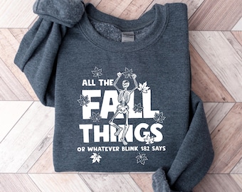 May include: A dark heather gray crewneck sweatshirt featuring a white graphic of a skeleton dancing among falling leaves. The text reads ALL THE FALL THINGS OR WHATEVER BLINK 182 SAYS in bold white lettering. The garment has long sleeves and a ribbed collar.