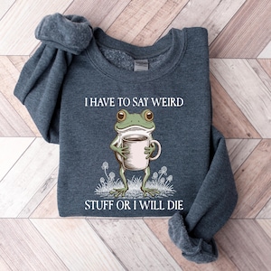 May include: A dark blue sweatshirt with the text "I HAVE TO SAY WEIRD STUFF OR I WILL DIE." The graphic features a green frog holding a coffee mug. The sweatshirt is folded on a light brown wooden surface.