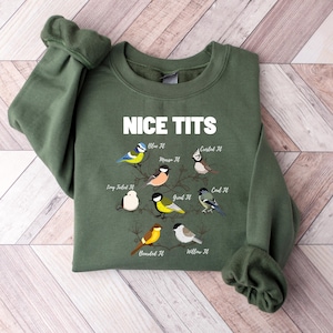 May include: An olive green sweatshirt with the text "NICE TITS" in white. Below the text are illustrations of various birds, each labeled with a bird species name. The sweatshirt is folded on a light brown wooden surface.