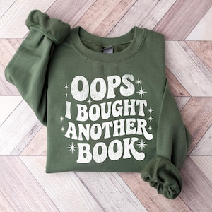 May include: An olive green sweatshirt with the text "OOPS I BOUGHT ANOTHER BOOK" in white, retro-style lettering. The sweatshirt is folded neatly on a light wood-grain surface. The sleeves are cuffed.