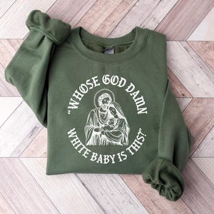 May include: An olive green sweatshirt with the text "WHOSE GOD DAMN WHITE BABY IS THIS?" in a circular design around a white illustration of Mary, Joseph, and baby Jesus. The sweatshirt is folded on a light wood surface.