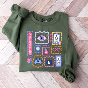 May include: An olive green sweatshirt featuring a colorful graphic design related to optometry. The design includes various framed images of eyes, eye charts, glasses, and other related symbols. The word "OPTOMETRY" is printed vertically in pink.