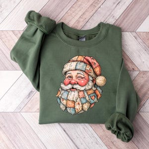 May include: An olive green sweatshirt featuring a whimsical Santa Claus graphic. The Santa has a rosy face, a white beard, and a patchwork hat and scarf in shades of red, orange, and blue. The sweatshirt has long sleeves.