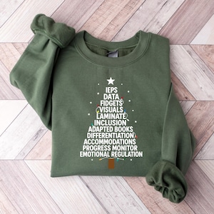 May include: An olive green sweatshirt with a Christmas tree design made of white text. The text includes words like "IEPS," "Data," "Fidgets," and "Inclusion." A star tops the tree, and string lights are incorporated into the design.