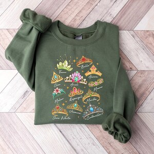 May include: Olive green sweatshirt featuring colorful illustrations of princess tiaras with names like Tiana, Ariel, and Cinderella. The design includes gold stars and script lettering. The sweatshirt has long sleeves and a crew neck.