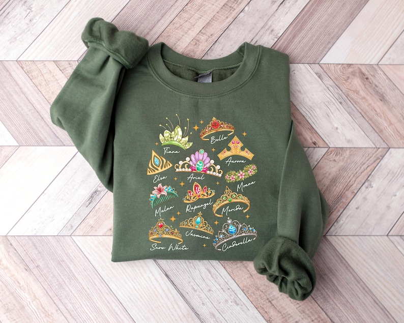 May include: Olive green sweatshirt featuring colorful illustrations of princess tiaras with names like Tiana, Ariel, and Cinderella. The design includes gold stars and script lettering. The sweatshirt has long sleeves and a crew neck.