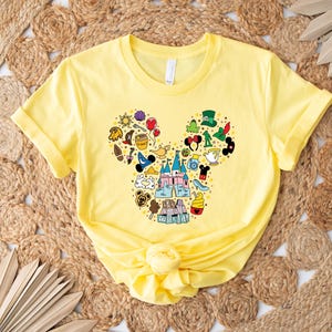 Puede incluir: Una camiseta amarilla con un dise&ntilde;o de cabeza de Mickey Mouse lleno de iconos coloridos con tem&aacute;tica de Disney. El dise&ntilde;o incluye un castillo, siluetas de personajes y otros elementos reconocibles. La camiseta est&aacute; hecha de un material suave y tiene mangas cortas.