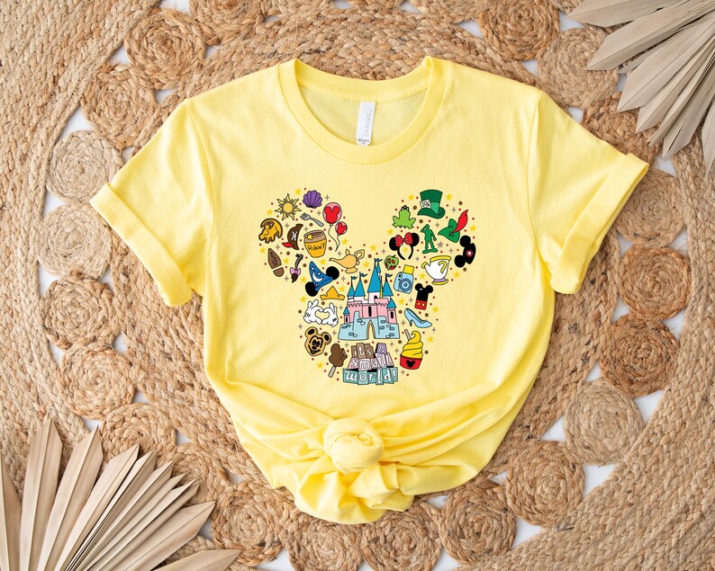 Puede incluir: Una camiseta amarilla con un dise&ntilde;o de cabeza de Mickey Mouse lleno de iconos coloridos con tem&aacute;tica de Disney. El dise&ntilde;o incluye un castillo, siluetas de personajes y otros elementos reconocibles. La camiseta est&aacute; hecha de un material suave y tiene mangas cortas.