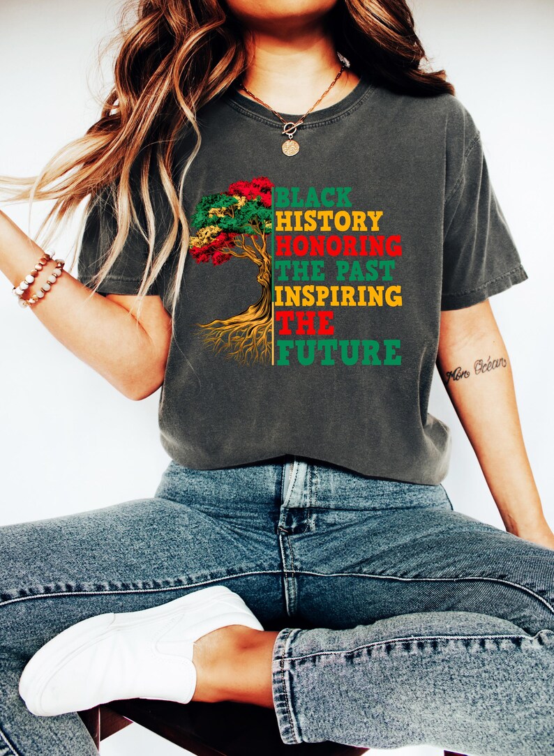 Pu&ograve; includere: T-shirt grigio scuro con la scritta "BLACK HISTORY HONORING THE PAST INSPIRING THE FUTURE" in rosso, verde e giallo. La maglietta presenta una grafica ad albero con foglie e radici rosse, verdi e gialle.