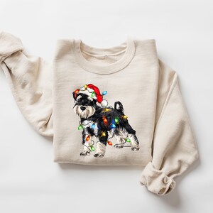 May include: A beige sweatshirt featuring a black and white miniature schnauzer wearing a red Santa hat and tangled in Christmas lights.