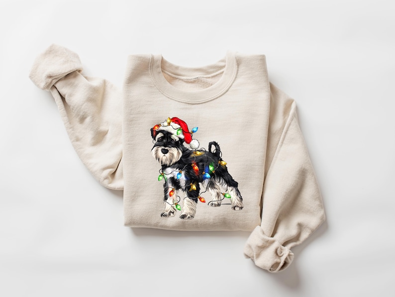 May include: A beige sweatshirt featuring a black and white miniature schnauzer wearing a red Santa hat and tangled in Christmas lights.