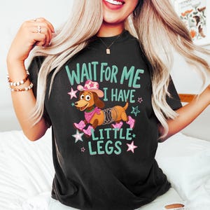 Puede incluir: Una camiseta negra con el gr&aacute;fico de un perro salchicha marr&oacute;n que lleva un sombrero de vaquero rosa, un pa&ntilde;uelo rosa y botas de vaquero rosas. El perro tiene un resorte met&aacute;lico por cuerpo. El texto dice WAIT FOR ME I HAVE LITTLE LEGS en letras verde azulado.