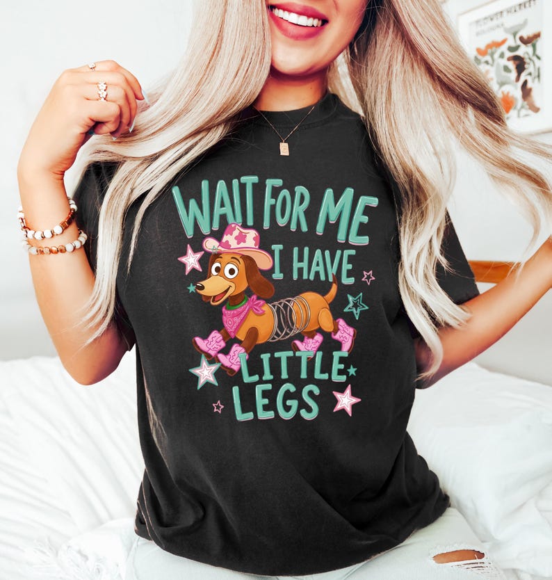 Puede incluir: Una camiseta negra con el gr&aacute;fico de un perro salchicha marr&oacute;n que lleva un sombrero de vaquero rosa, un pa&ntilde;uelo rosa y botas de vaquero rosas. El perro tiene un resorte met&aacute;lico por cuerpo. El texto dice WAIT FOR ME I HAVE LITTLE LEGS en letras verde azulado.