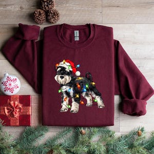 May include: A maroon long-sleeved sweatshirt featuring a black and white miniature schnauzer wearing a Santa hat and tangled in Christmas lights. The dog is centrally positioned on the sweatshirt.