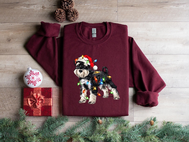 May include: A maroon long-sleeved sweatshirt featuring a black and white miniature schnauzer wearing a Santa hat and tangled in Christmas lights. The dog is centrally positioned on the sweatshirt.