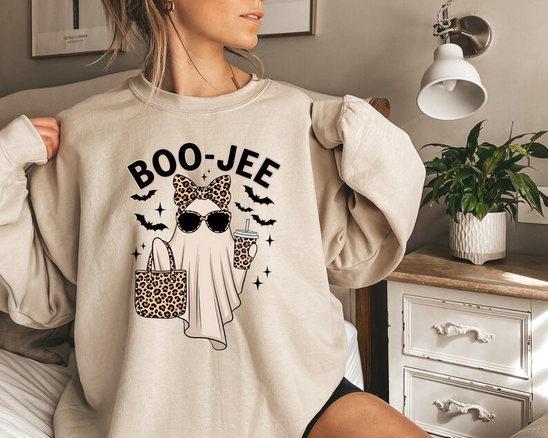 May include: Beige sweatshirt with a Halloween graphic. The design shows a ghost wearing sunglasses and a leopard print bow, holding a leopard print tote bag and a drink. The word "BOO-JEE" is above the ghost, with bats and stars around it.