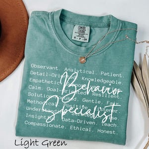 May include: Light green t-shirt with the words "Behavior Specialist" in white script. The shirt also lists positive attributes. A gold necklace with a pendant rests on the shirt.