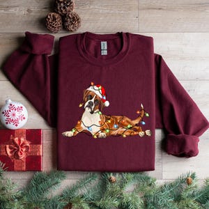 May include: A maroon long-sleeved sweatshirt with a Christmas-themed design featuring a brown and white boxer dog wearing a Santa hat and wrapped in colorful Christmas lights.