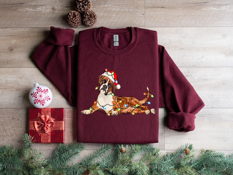 May include: A maroon long-sleeved sweatshirt with a Christmas-themed design featuring a brown and white boxer dog wearing a Santa hat and wrapped in colorful Christmas lights.