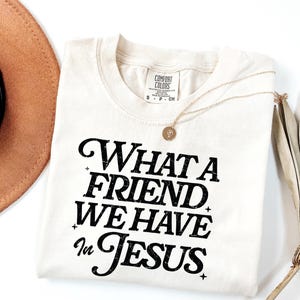 K&ouml;nnte beinhalten: Ein cremefarbenes T-Shirt mit schwarzem Text, der "What a Friend We Have in Jesus" lautet.