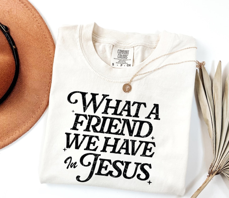 K&ouml;nnte beinhalten: Ein cremefarbenes T-Shirt mit schwarzem Text, der "What a Friend We Have in Jesus" lautet.