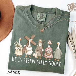 May include: Moss green t-shirt with a graphic of cartoon geese dressed in biblical attire, holding a cross, flowers, and a basket of eggs. The text "HE IS RISEN SILLY GOOSE" is printed below the geese. A gold necklace is draped on the shirt.