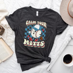 May include: A dark gray t-shirt with a retro baseball graphic and the text "Calm Your Mitts".
