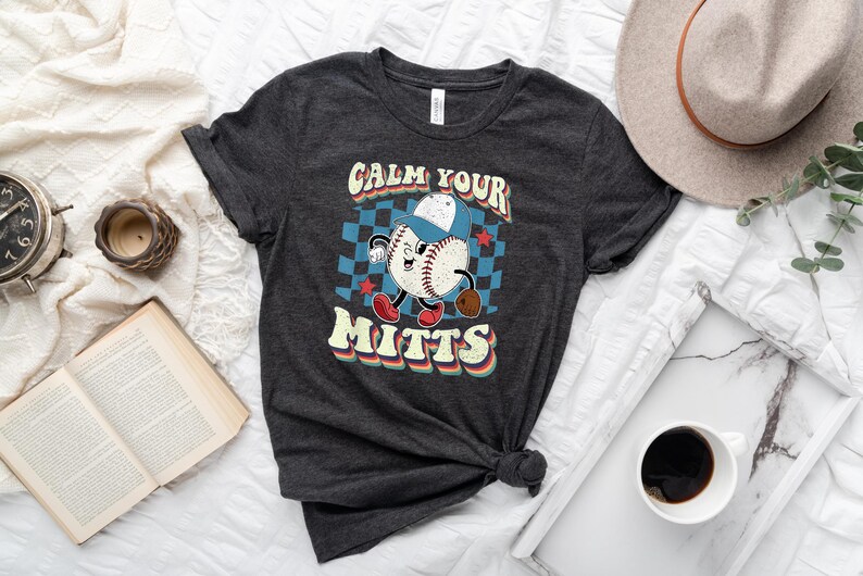 May include: A dark gray t-shirt with a retro baseball graphic and the text "Calm Your Mitts".