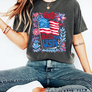 May include: A charcoal grey short-sleeve t-shirt featuring a graphic print of an American flag with the text GOD BLESS THE USA in red and blue. The design is surrounded by colorful floral and leaf illustrations in shades of red, white, and blue.