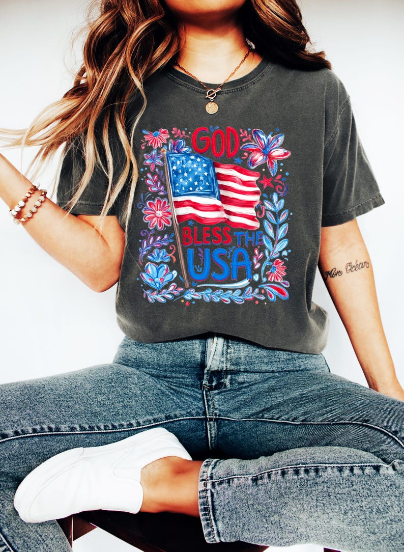 May include: A charcoal grey short-sleeve t-shirt featuring a graphic print of an American flag with the text GOD BLESS THE USA in red and blue. The design is surrounded by colorful floral and leaf illustrations in shades of red, white, and blue.