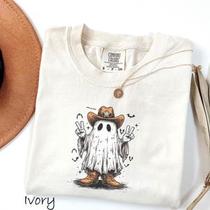 Pu&ograve; includere: T-shirt color avorio con un fantasma dei cartoni animati che indossa un cappello da cowboy e stivali, che fa il segno della pace. La grafica include pipistrelli e stelle. Sono visibili anche una collana d'oro e un cappello marrone.