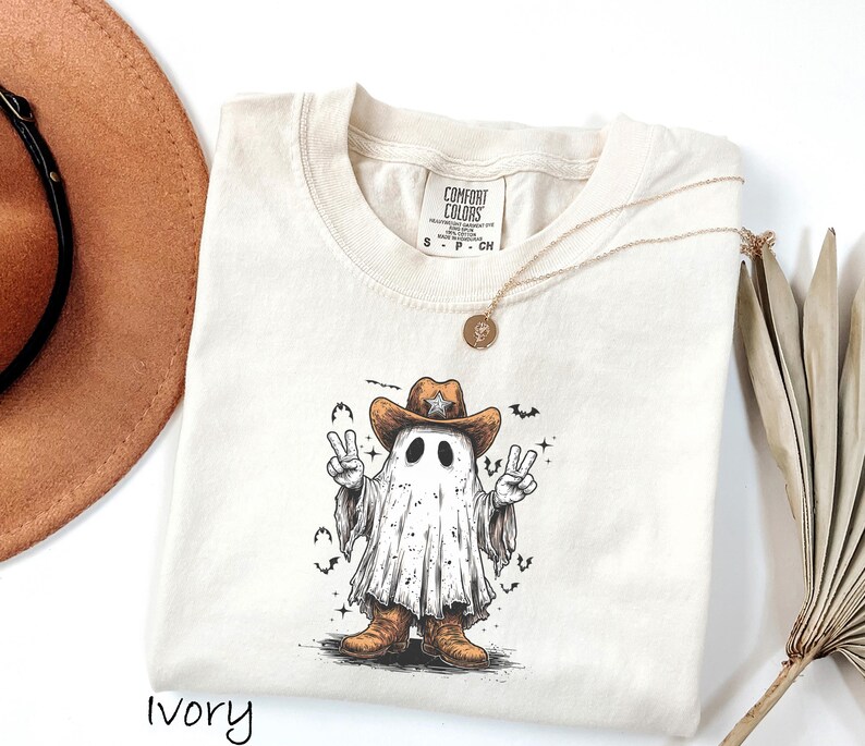 Pu&ograve; includere: T-shirt color avorio con un fantasma dei cartoni animati che indossa un cappello da cowboy e stivali, che fa il segno della pace. La grafica include pipistrelli e stelle. Sono visibili anche una collana d'oro e un cappello marrone.
