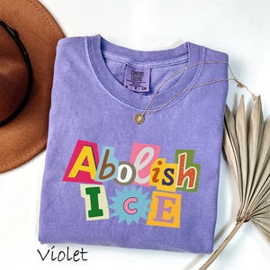 May include: A violet-colored crew neck t-shirt with the words "Abolish ICE" in a colorful, cut-out style design. The shirt is made of a soft material and has a tag that says "Comfort Colors". A gold necklace is draped across the shirt.
