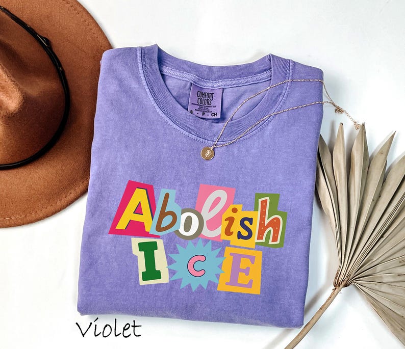 May include: A violet-colored crew neck t-shirt with the words "Abolish ICE" in a colorful, cut-out style design. The shirt is made of a soft material and has a tag that says "Comfort Colors". A gold necklace is draped across the shirt.