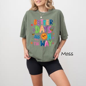 May include: Moss green t-shirt with the text "Better Days Are On The Way" in colorful, playful lettering. The design includes a smiling sun, bees, and hearts. The shirt is paired with black shorts.