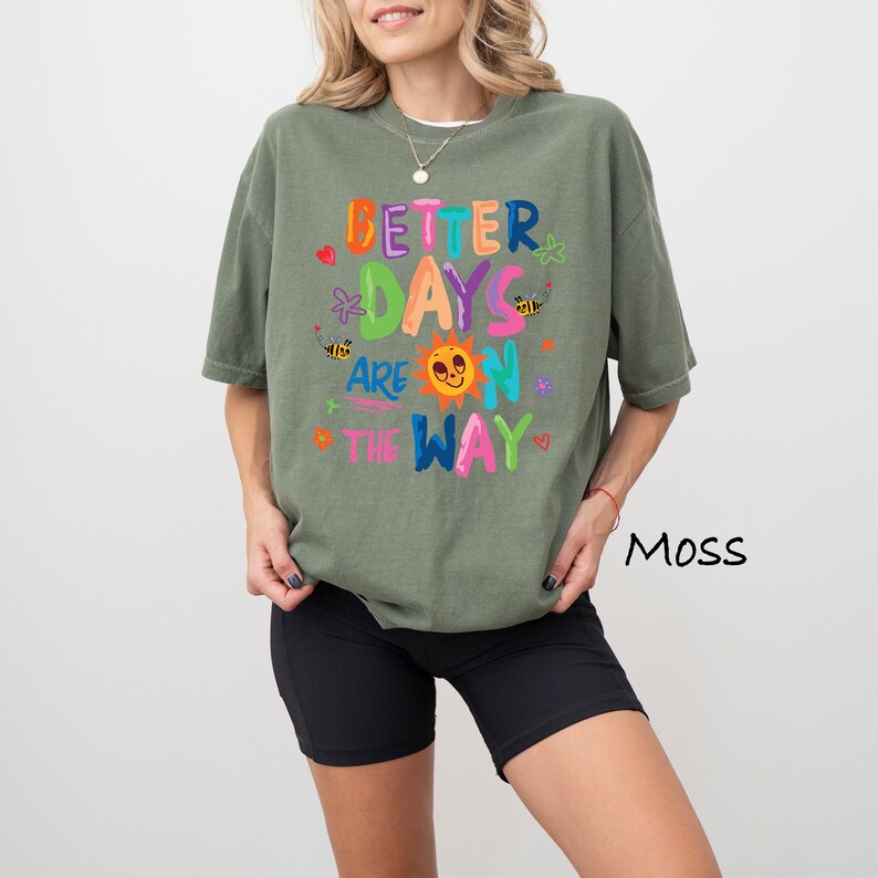 May include: Moss green t-shirt with the text "Better Days Are On The Way" in colorful, playful lettering. The design includes a smiling sun, bees, and hearts. The shirt is paired with black shorts.