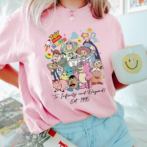 May include: A light pink t-shirt featuring a colorful graphic of Toy Story characters. The design includes the text "Toy Story Land" and "To Infinity and Beyond! Est. 1995". The shirt is paired with light blue shorts.