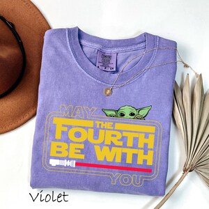 May include: A violet Comfort Colors t-shirt featuring a yellow graphic that reads "MAY THE FOURTH BE WITH YOU" with a green character peeking over the top. A red lightsaber is at the bottom of the graphic. A gold necklace and a brown hat are also in the image.