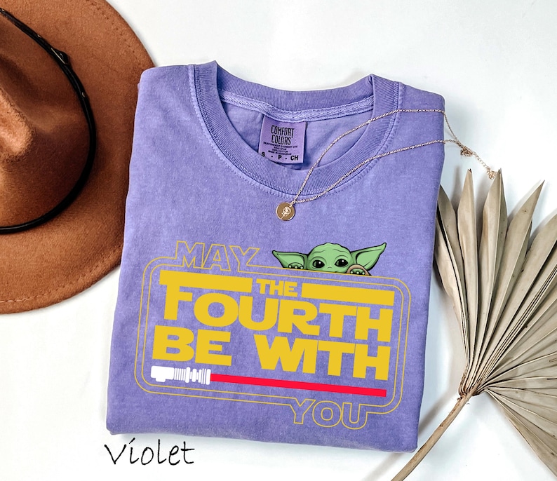 May include: A violet Comfort Colors t-shirt featuring a yellow graphic that reads "MAY THE FOURTH BE WITH YOU" with a green character peeking over the top. A red lightsaber is at the bottom of the graphic. A gold necklace and a brown hat are also in the image.
