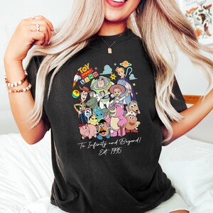 May include: Black t-shirt featuring a colorful graphic of Toy Story characters, including Woody, Buzz Lightyear, and others. The shirt has the text "Toy Story Land" at the top and "To Infinity and Beyond! Est. 1995" at the bottom.