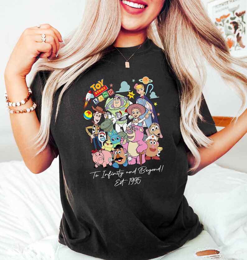 May include: Black t-shirt featuring a colorful graphic of Toy Story characters, including Woody, Buzz Lightyear, and others. The shirt has the text "Toy Story Land" at the top and "To Infinity and Beyond! Est. 1995" at the bottom.