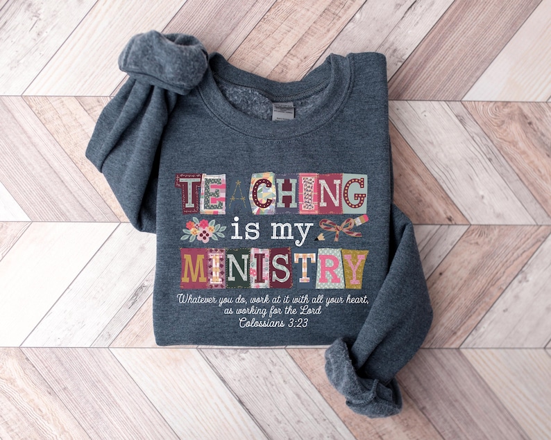 May include: A dark gray sweatshirt with the words "TEACHING is my MINISTRY" in colorful, block letters. Floral and bow accents are included in the design. The text "Whatever you do, work at it with all your heart, as working for the Lord Colossians 3:23" is also printed on the shirt.