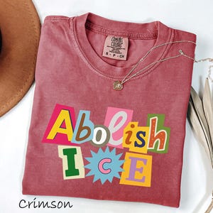 May include: Crimson-colored t-shirt with the words "Abolish ICE" in a colorful, cut-out style design. The shirt has a crew neck and short sleeves. A silver necklace with a small pendant is draped across the shirt.