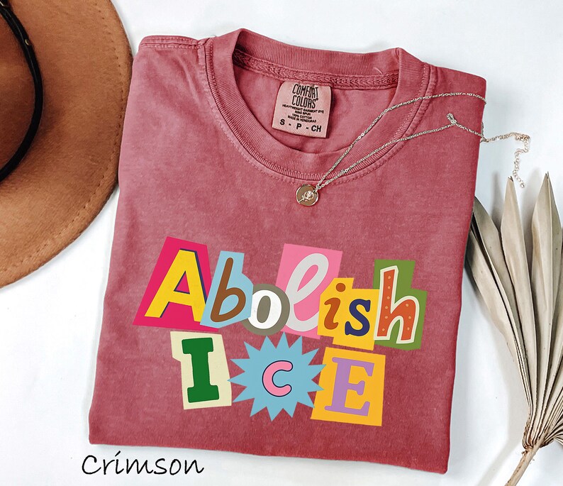 May include: Crimson-colored t-shirt with the words "Abolish ICE" in a colorful, cut-out style design. The shirt has a crew neck and short sleeves. A silver necklace with a small pendant is draped across the shirt.
