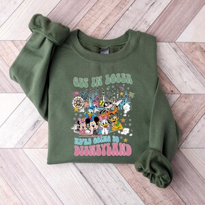 May include: Olive green sweatshirt with the text "GET IN LOSER WE'RE GOING TO DISNEYLAND" in a retro font. The graphic features cartoon characters and a colorful illustration of a theme park.